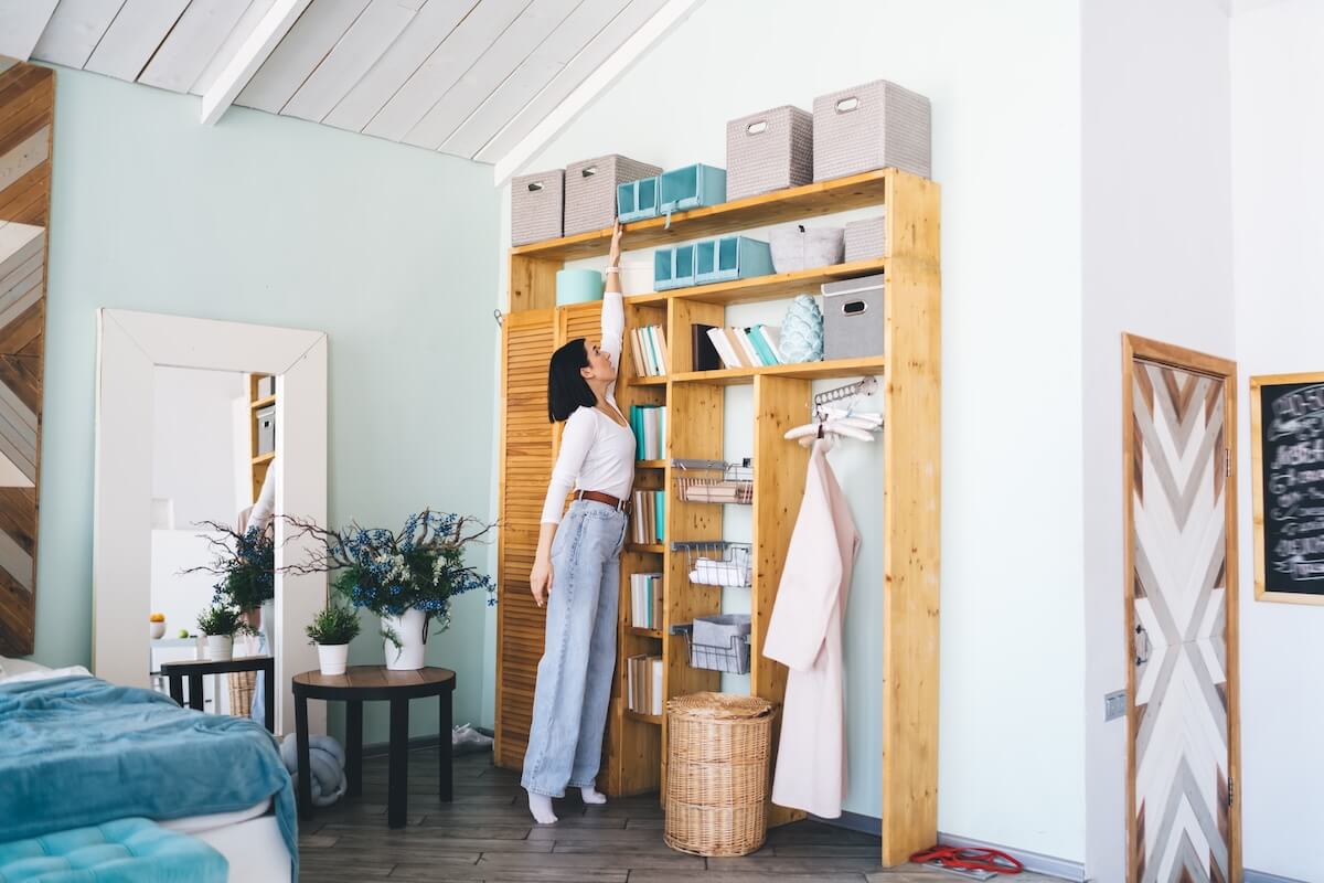 Shutterstock: Full body of young female in casual clothes reaching for blue storage box on top shelf of wooden wardrobe in modern apartment