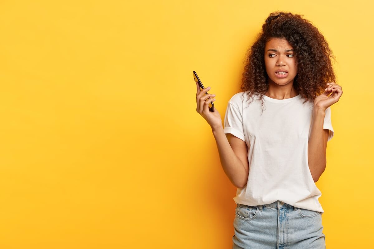 Shutterstock: Beautiful female with frizzy Afro hairstyle, looks in displeasure at cell phone, receives call from former boyfriend, holds mobile phone on distance, ignores communication, afraids of something