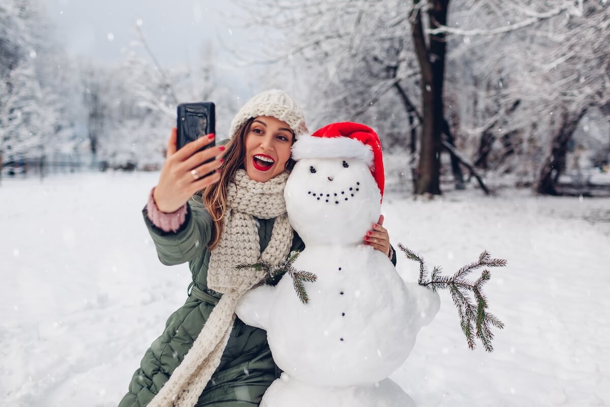 Shutterstock: Happy woman takes selfie by snowman in Santa hat using phone outdoors in snowy winter park. Christmas festive season. Fun activities