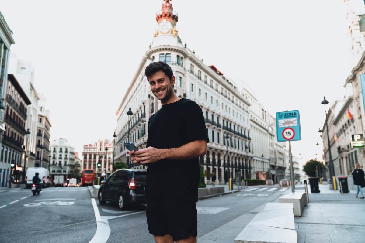 Shutterstock: Cheerful young man in black outfit smiling while checking smartphone app on city street, historic architecture in background, relaxed morning vibes in busy European downtown. Male using mobile phone