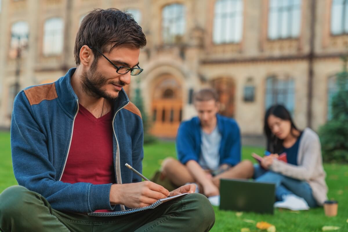 Shutterstock: Young concentered male student man guy noting doing homework task sitting on the grass while two college students classmates using laptop studding in background at university campus lawn.