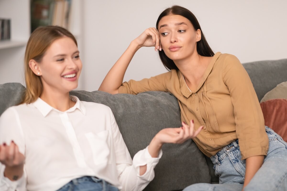 Shutterstock: Envy In Friendship. Jealous Lady Listening To Her Cheerful Friend Talking And Bragging About Her Great Life Sitting On Couch At Home, Rolling Eyes During Conversation. Selective Focus