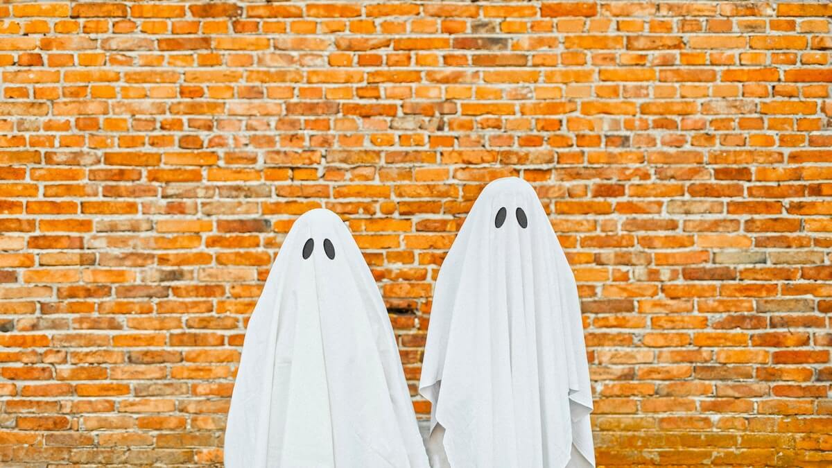 Shutterstock: Two people dressed as ghosts with white sheets and black eye holes stand in front of a vibrant brick wall, giving a playful nod to classic Halloween costumes.