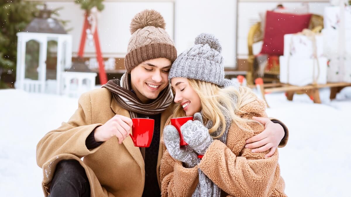Shutterstock: A young couple sits together in a snowy outdoor setting, sharing warm drinks from red mugs. They are bundled in cozy winter attire, radiating joy and warmth against the cold backdrop.