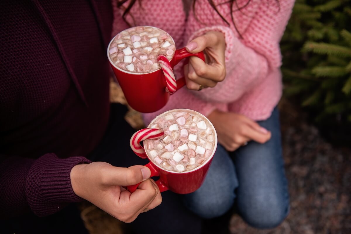 Shutterstock: Close-up of hands of young couple holding red cups with hot cocoa, marshmallows, striped red, white candy cane. Man and woman are having fun at Christmas tree market. Faceless view. Copy space