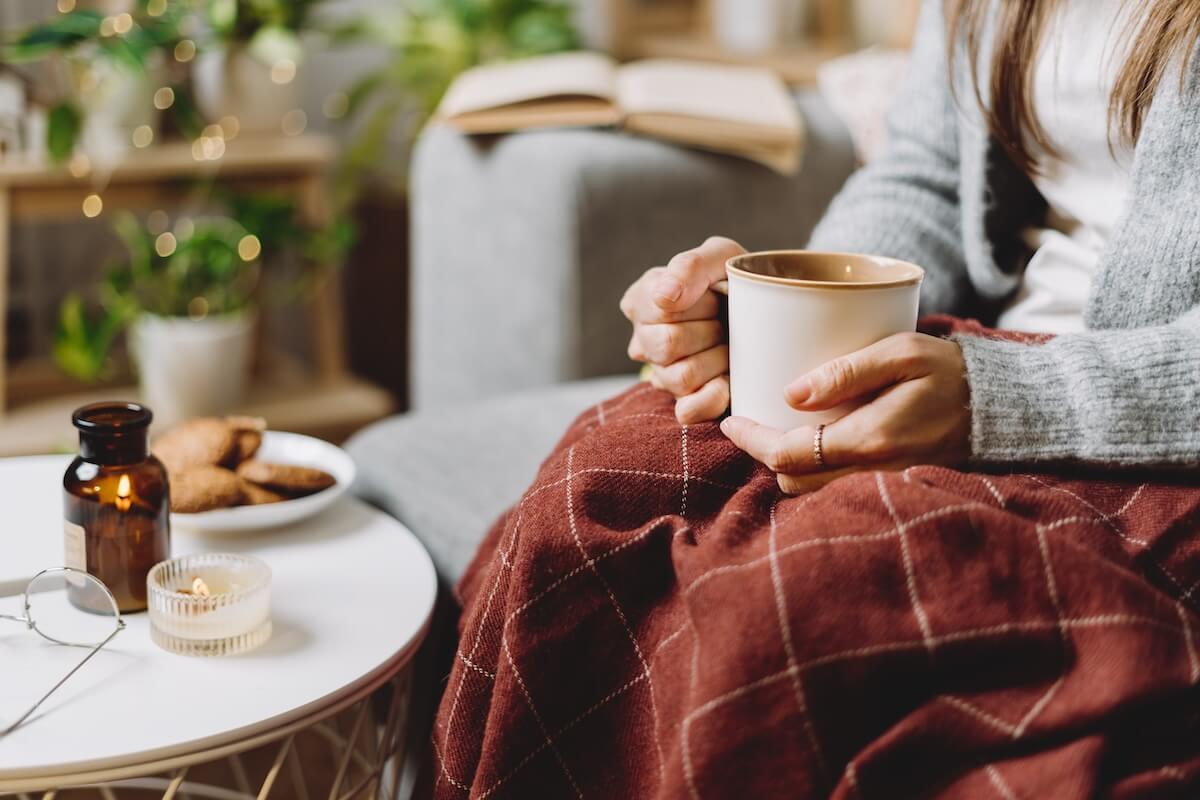 Shutterstock: Cozy woman legs in knitted winter warm sweater and checkered plaid drinking hot cocoa or coffee in mug, sitting on couch at home. Autumn vibes with candle, decor, cookies and indoor plants