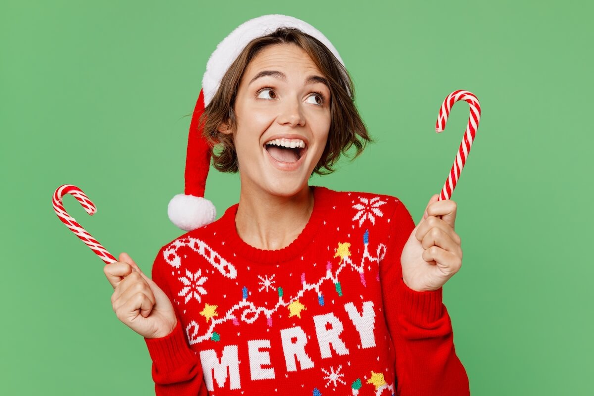 Shutterstock: Merry young fun woman wears knitted xmas sweater Santa hat posing hold in hand candy cane look aside isolated on plain pastel light green background. Happy New Year 2023 celebration holiday concept