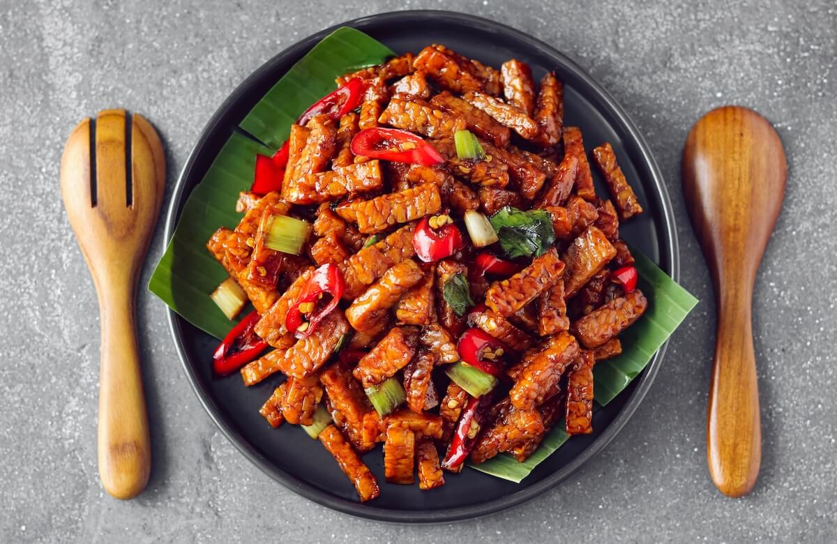 Shutterstock: Tempeh or Tempe Orek. A dish featuring stir-fried tempeh strips, coated in a glossy, savory sweet sauce, and contrasted with bright red chilies and fresh green spring onions.