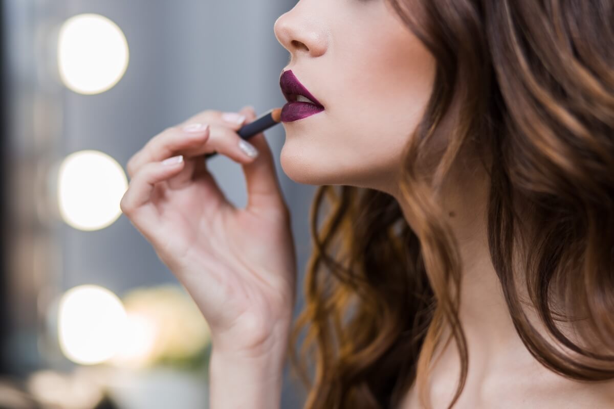 Shutterstock: Beautiful brunette girl smears on bright Burgundy lip pencil, close-up, horizontal, cosmetics