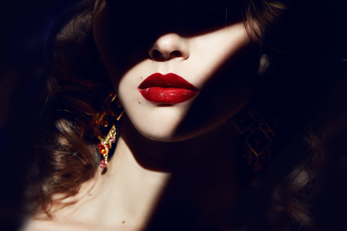 Shutterstock: Closeup of the face belong to beautiful young sexy blonde girl with curly hair pure snow white skin and bright makeup red lips, red lipstick, long earrings in dark shadows of palm leaves