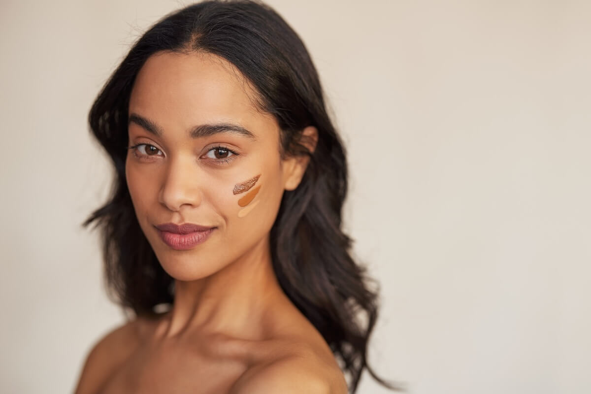 Shutterstock: Portrait of young latin hispanic woman applying foundation swatches of various tone on face while looking at camera. Beautiful mixed race makeup artist with swatches of foundation cream on cheek.