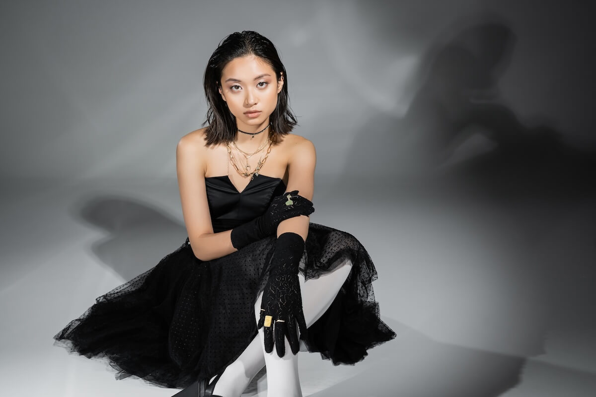 Shutterstock: stylish asian young woman with short hair sitting in black strapless dress with tulle skirt, white tights and gloves while looking at camera on grey background, wet hairstyle, golden necklaces