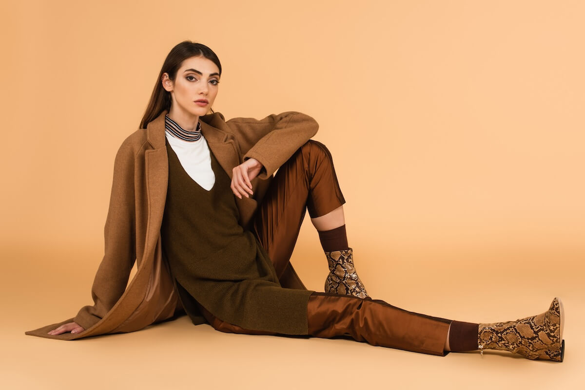 Shutterstock: young woman in brown coat, satin pants and leather shoes sitting on beige background