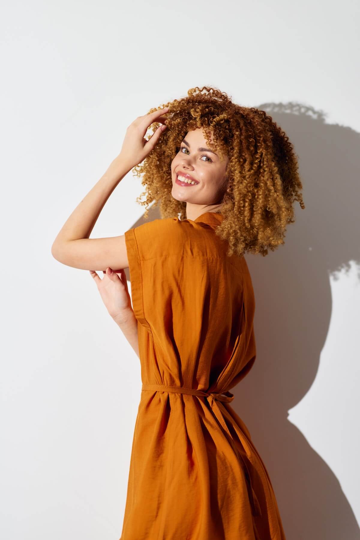 Shutterstock: Stylish woman with curly hair wearing an orange dress against a plain white background, showcasing a cheerful and confident pose with a playful smile