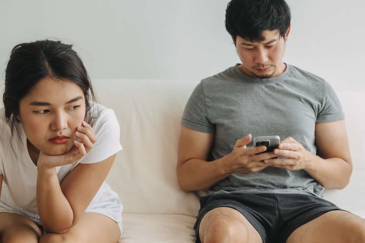 Shutterstock: Asian Thai man using mobile phone and ignoring girlfriend, woman sad, both sitting on white sofa in room apartment, married life concept.
