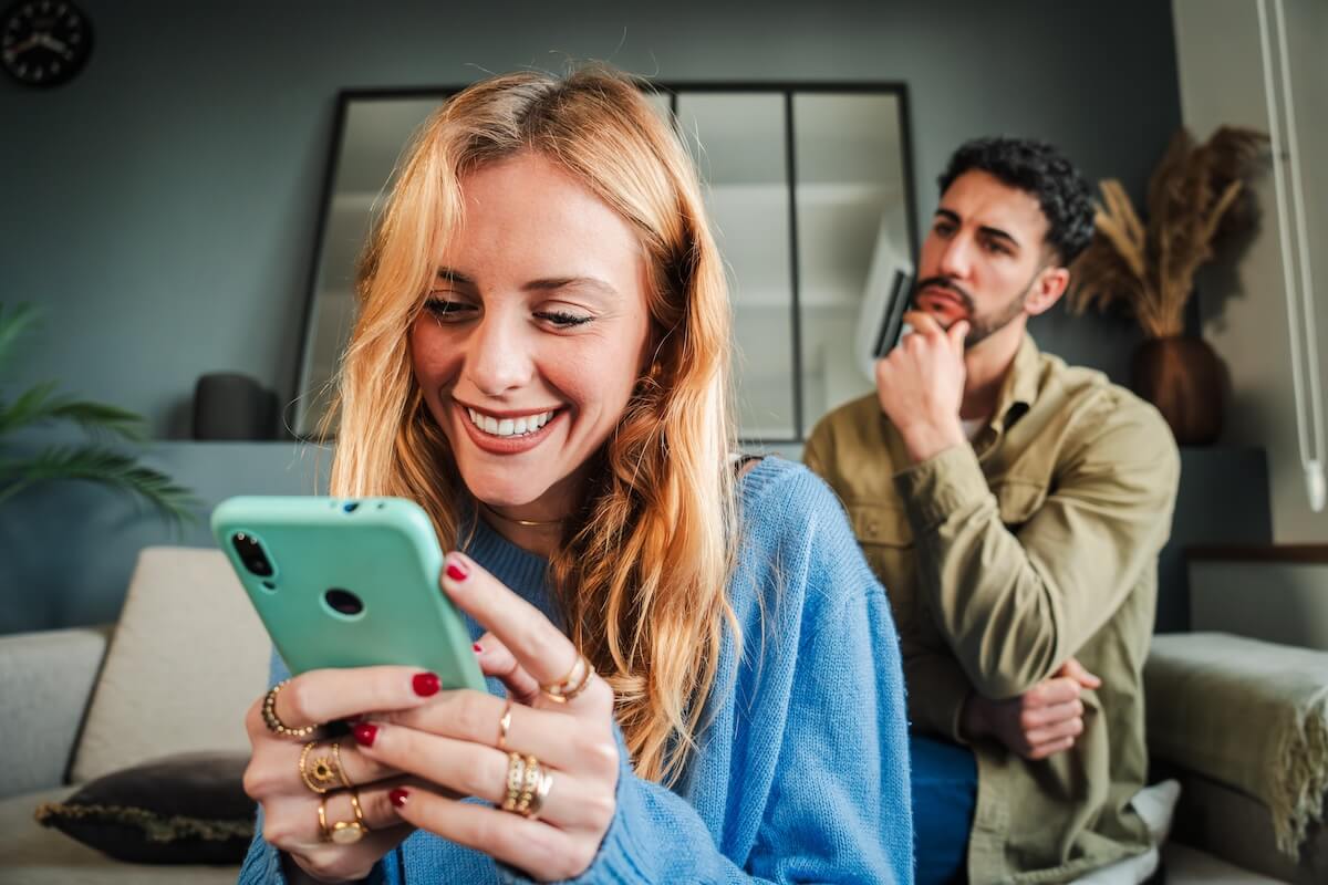 Shutterstock: Woman secretly texting on her phone to deceive her husband, while he peeks over her shoulder and reads her messages. The boyfriend, feeling angry and upset, watches his girlfriend as she pays him no