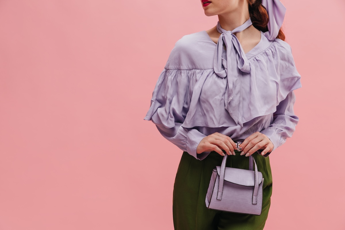 Shutterstock: Close-up of below neck stylish young caucasian woman on pink background. Red-haired model with red lips in lilac blouse, green pants and woman's handbag. Beauty and fashion, concept