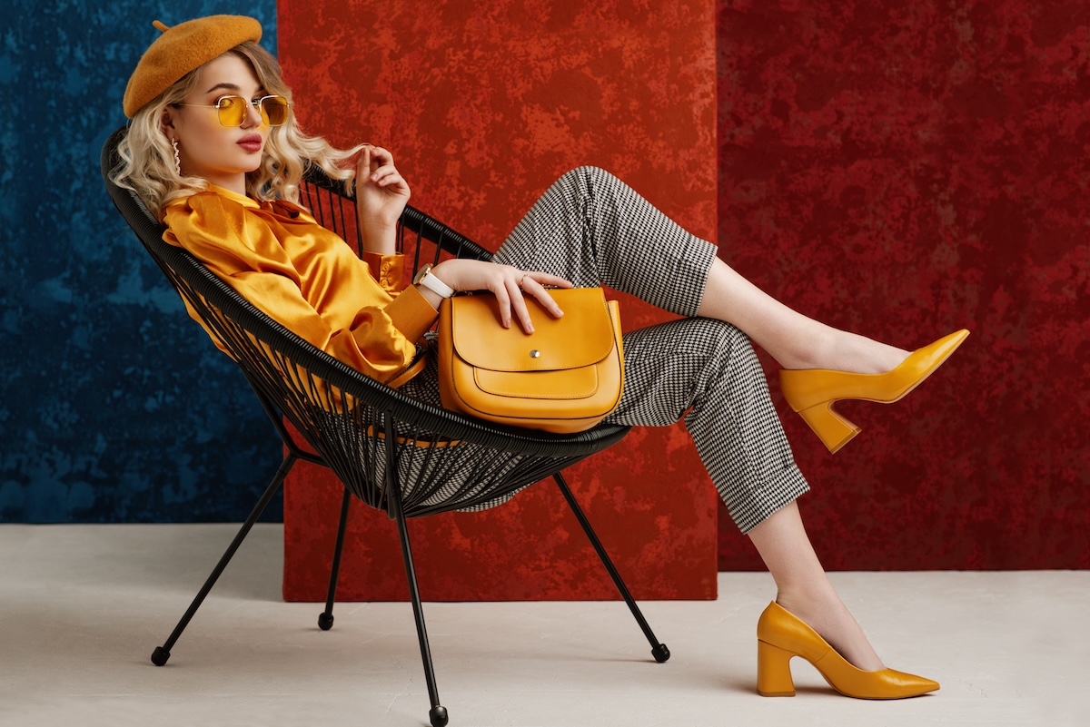Shutterstock: Full-length studio fashion portrait of elegant woman wearing yellow color sunglasses, beret, silk blouse, houndstooth printed trousers, pointed toe shoes, posing on chair, holding stylish leather bag