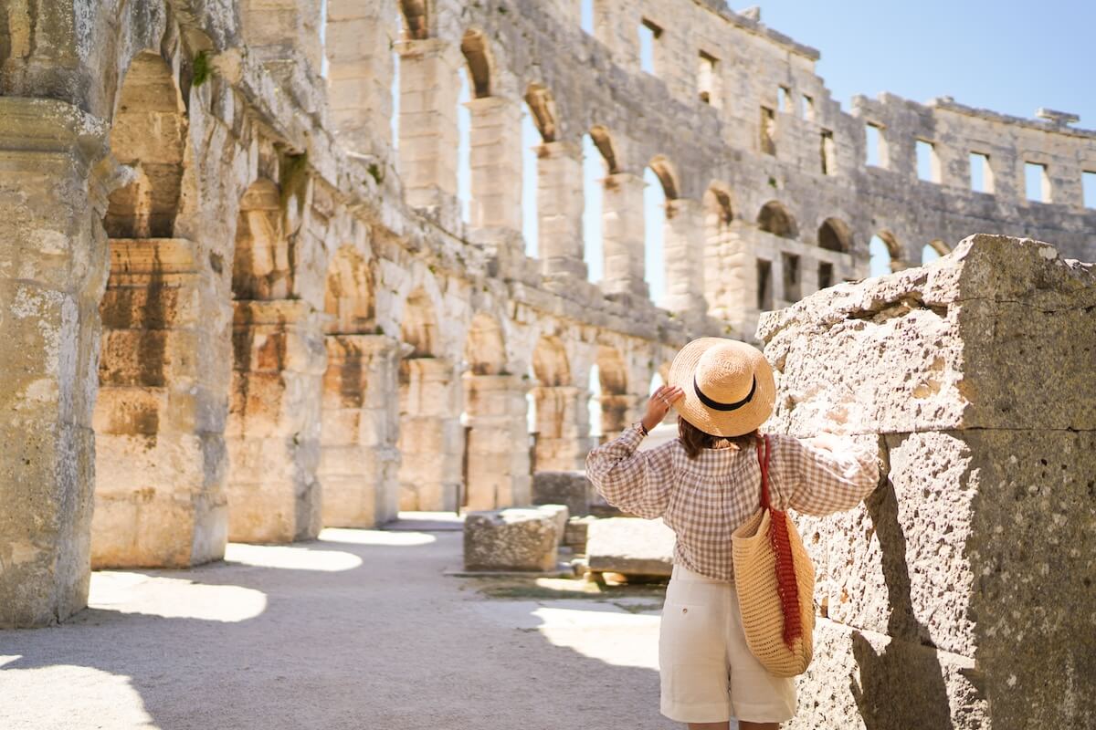 Shutterstock: woman in hat stands and looking on Coliseum in Pula Croatia. travel concept.Young woman enjoy summer Italian vacation in Europe.