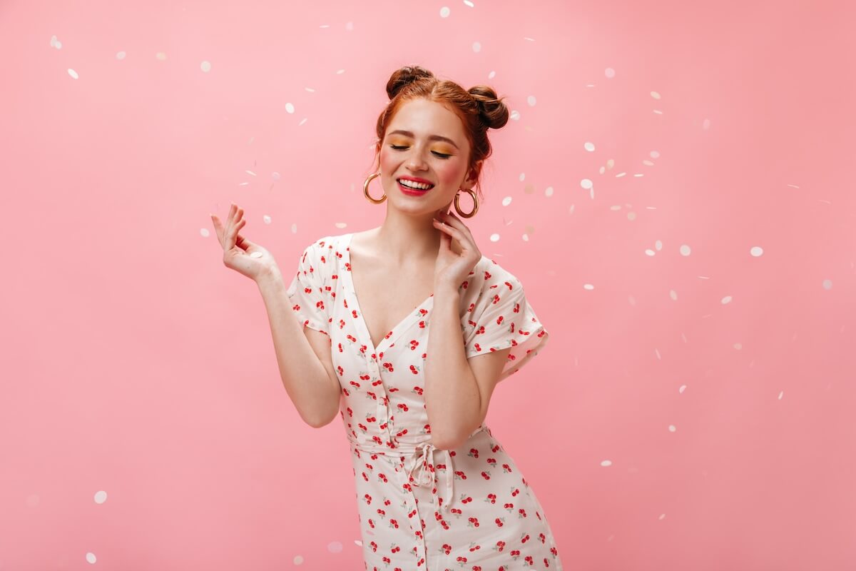 Shutterstock: Charming lady in white dress with cherries smiles affably. Portrait of redhead girl in massive earrings on pink background