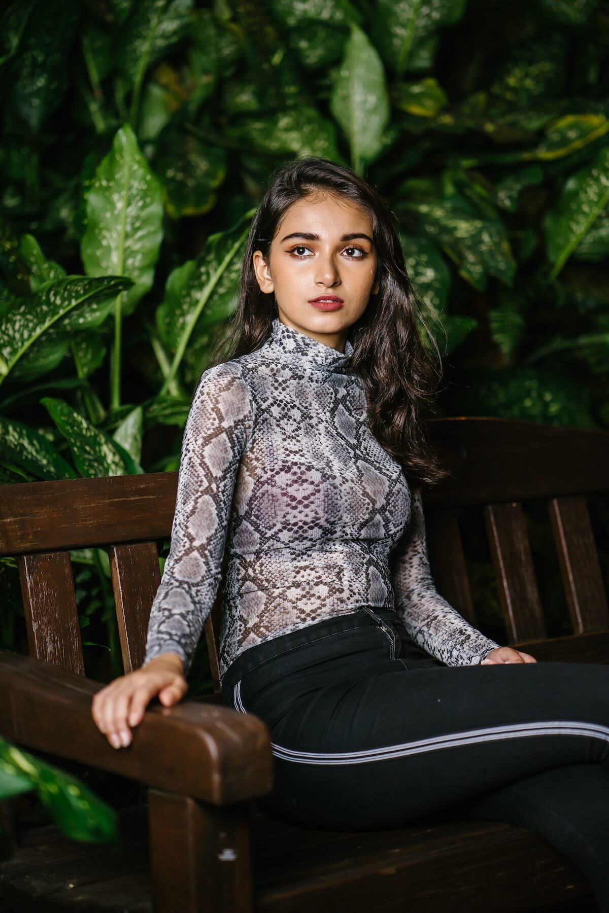 Shutterstock: Fashion portrait of a young, beautiful and elegant Indian Asian woman model in a snakeskin top and black pants sitting on a bench in a park in the evening. She is elegant and poised.