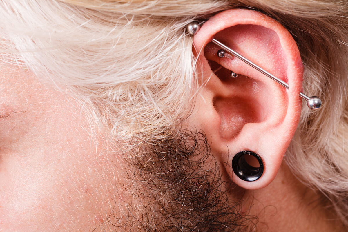 Man with industrial piercing and gauge earring: Shutterstock. Stretched lobe piercing, grunge concept. Pierced man ear with black plug tunnel. industrial and rook