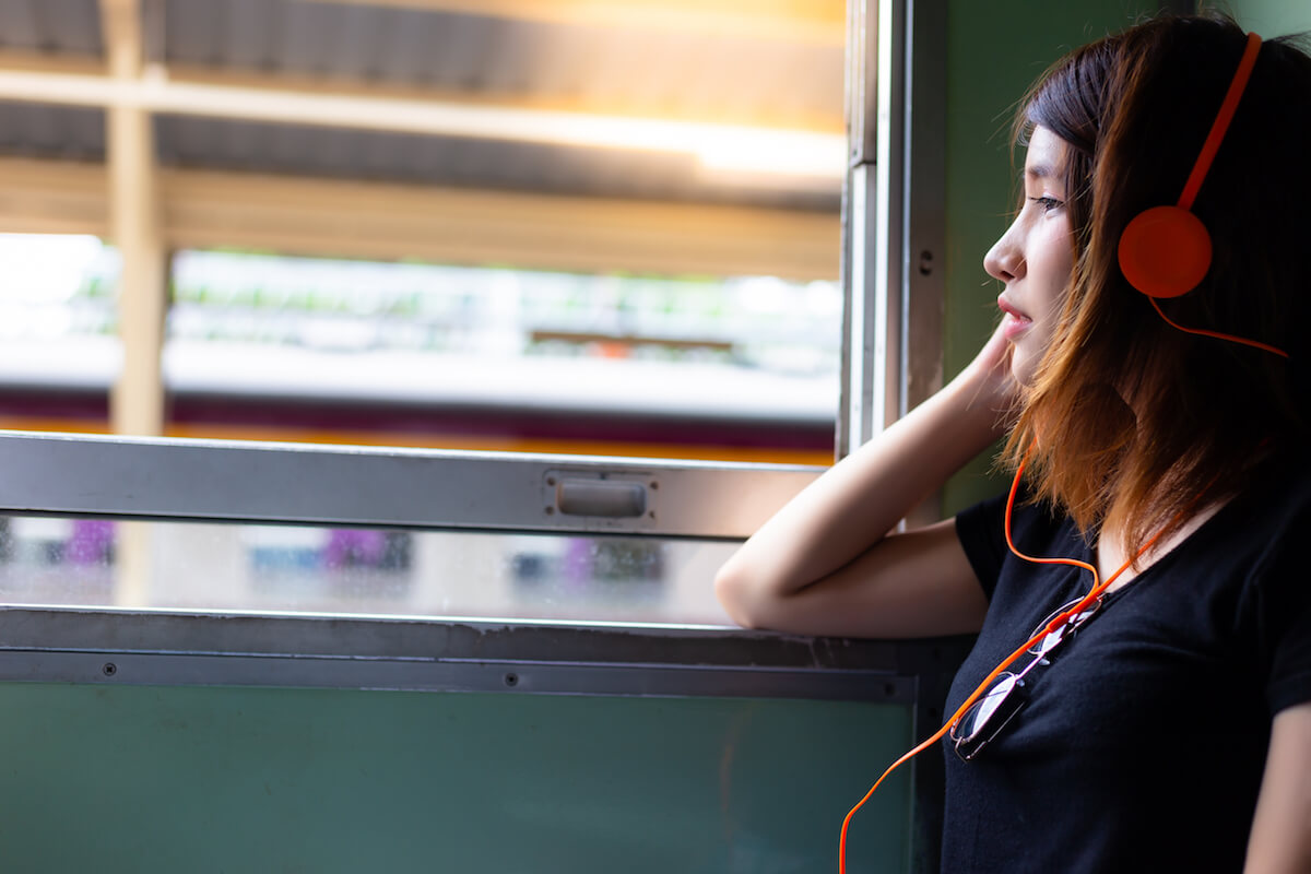 Woman on train sad and listening to headphones: Shutterstock. Portrait charming beautiful lovely woman. Attractive girl is listening sad music that make her feeling lonely, sad and miss someone. Pretty girl travel alone and look outside a retro train. copy space