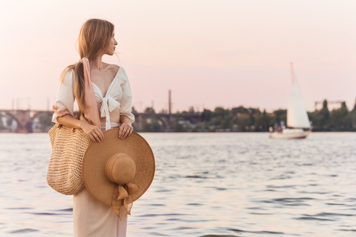 Shutterstock: A stylish young woman in a white blouse and skirt, holding a straw hat and woven bag, standing by the water with a peaceful expression at sunset. Ideal for lifestyle, fashion, or travel themes.