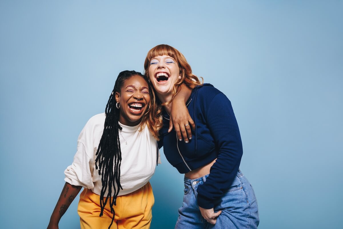 Shutterstock: Best friends laughing and having a good time together in a studio. Happy young women enjoying themselves while standing against a blue background. Two vibrant female friends making memories.