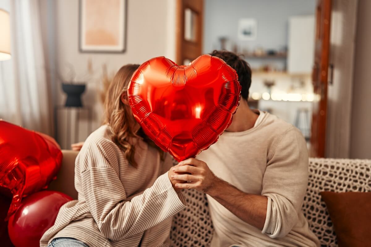 Shutterstock: Happy Valentine's Day. Young couple in love holding a heart-shaped balloon, covering themselves with it while kissing, sitting on the sofa in the living room at home. Romantic evening together.