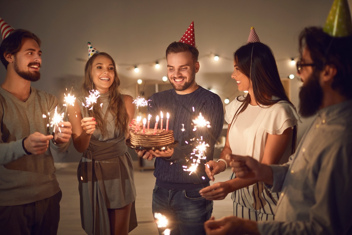 Shutterstock: Group of cheerful young people in cone party hats with lit sparklers in hands wishing happy birthday to their friend whos about to blow candles on his birthday cake during celebration at home