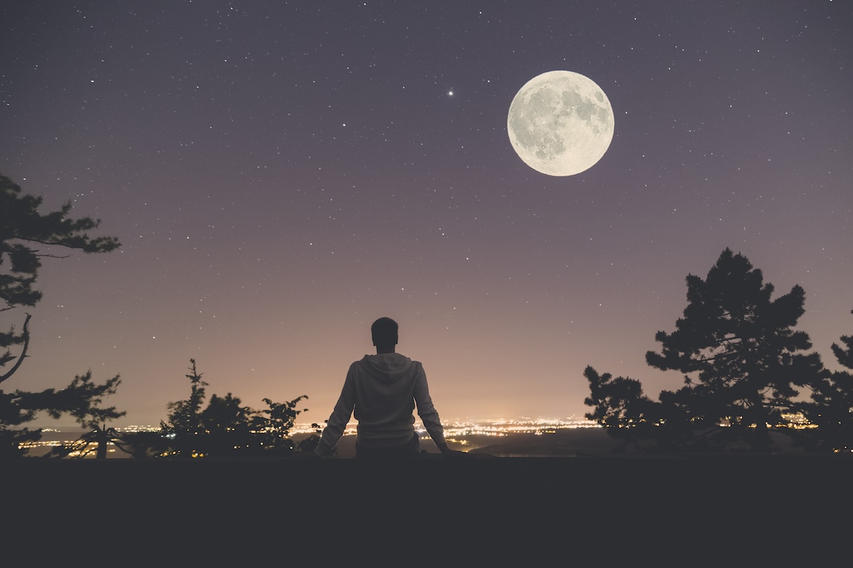 Shutterstock: Young man sitting on the wall at night. City lights, moon and stars in the background.