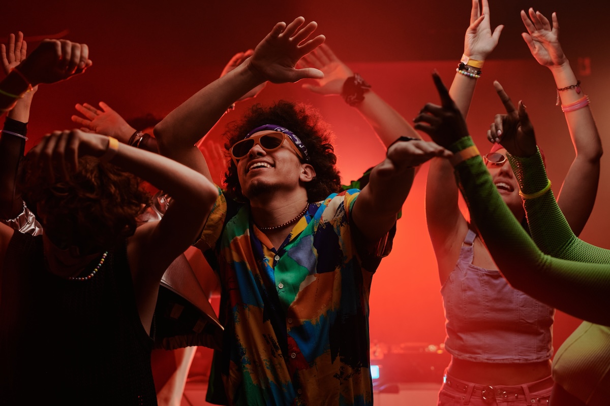 Shutterstock: Young excited man in stylish apparel and trendy sunglasses keeping his arms raised while dancing at disco party among his friends