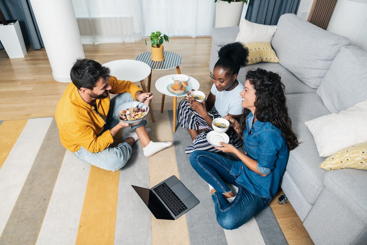 Shutterstock: Three young carefree friends two women and one man sitting in the apartment on the floor listening to music on a laptop eating and having fun for a free weekend without worries