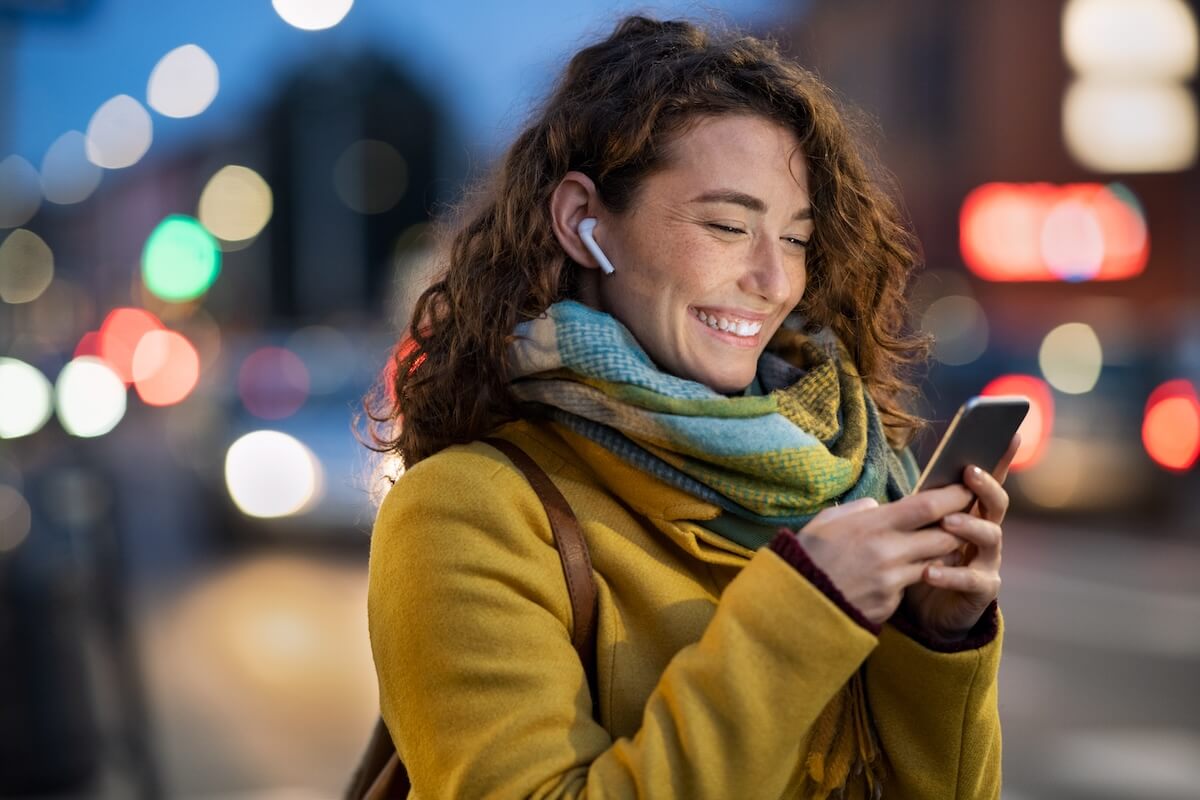Shutterstock: Beautiful woman using mobile phone while listening voice note with wireless earbuds. Smiling girl changing songs from smart phone in a city street. Happy smiling woman chatting on smartphone in winter