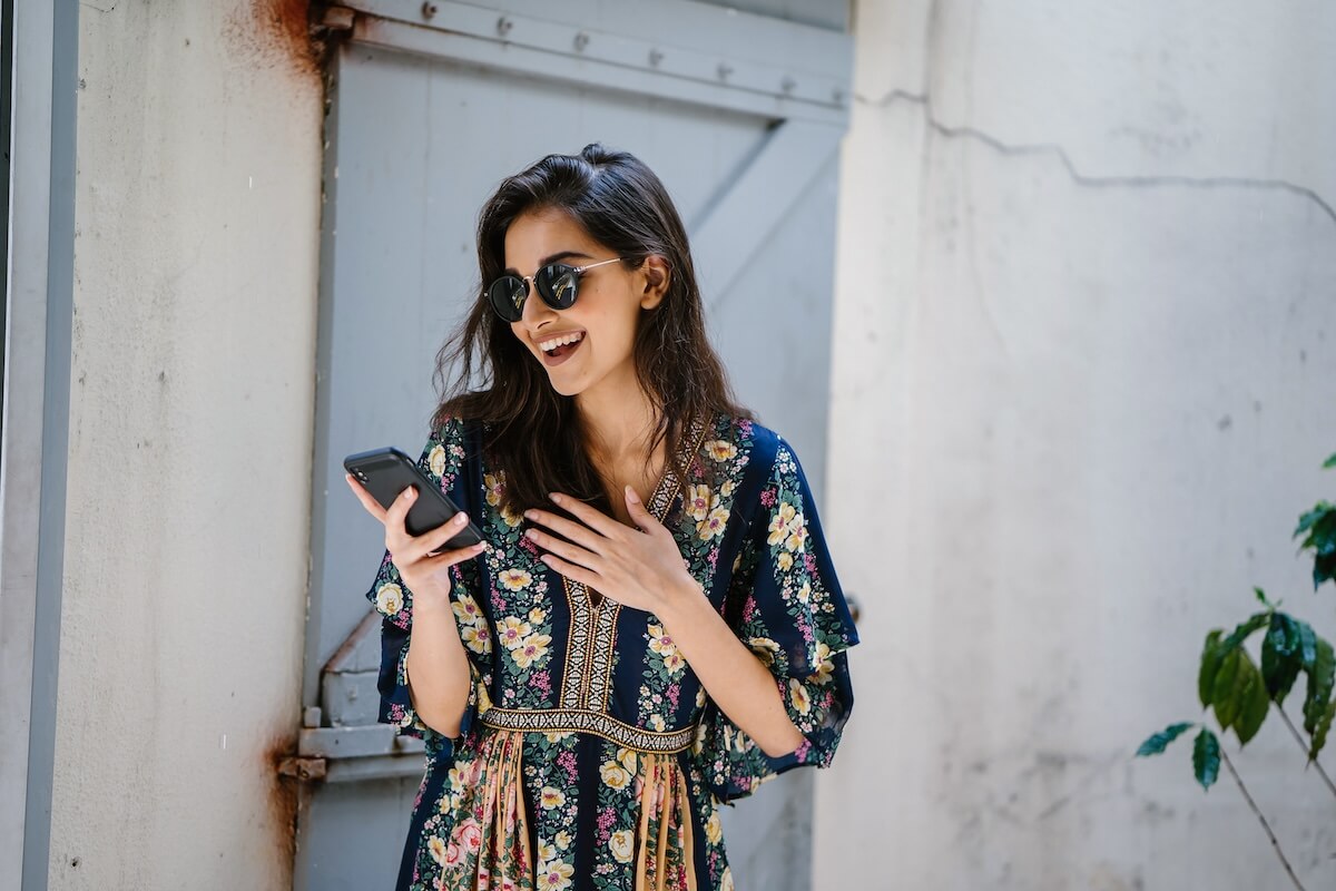 Shutterstock: Portrait of a young, attractive and beautiful Indian Asian woman wearing a dress and sunglasses with her smartphone. She looks surprised and delighted and is smiling as she looks at her phone.