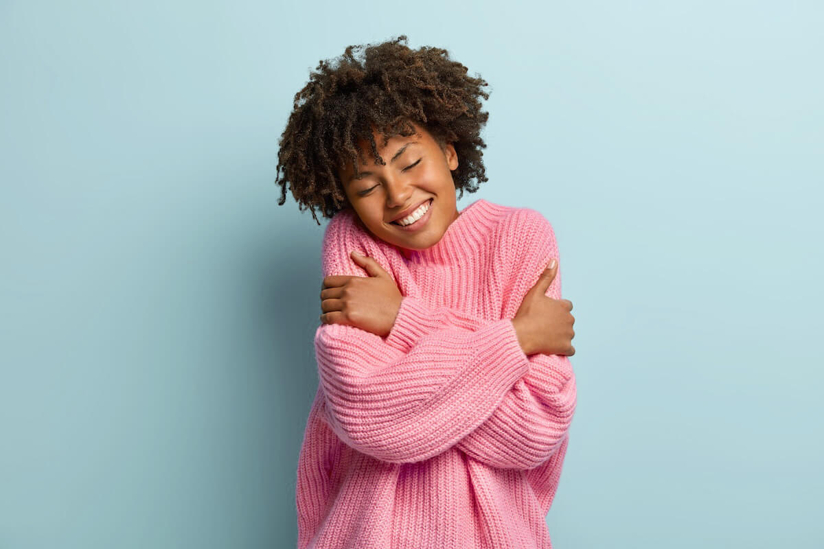 Shutterstock: Love yourself concept. Photo of lovely smiling woman embraces herself, has high self esteem, closes eyes from enjoyment, likes her new comfortable soft pink sweater, tilts head, stands indoor