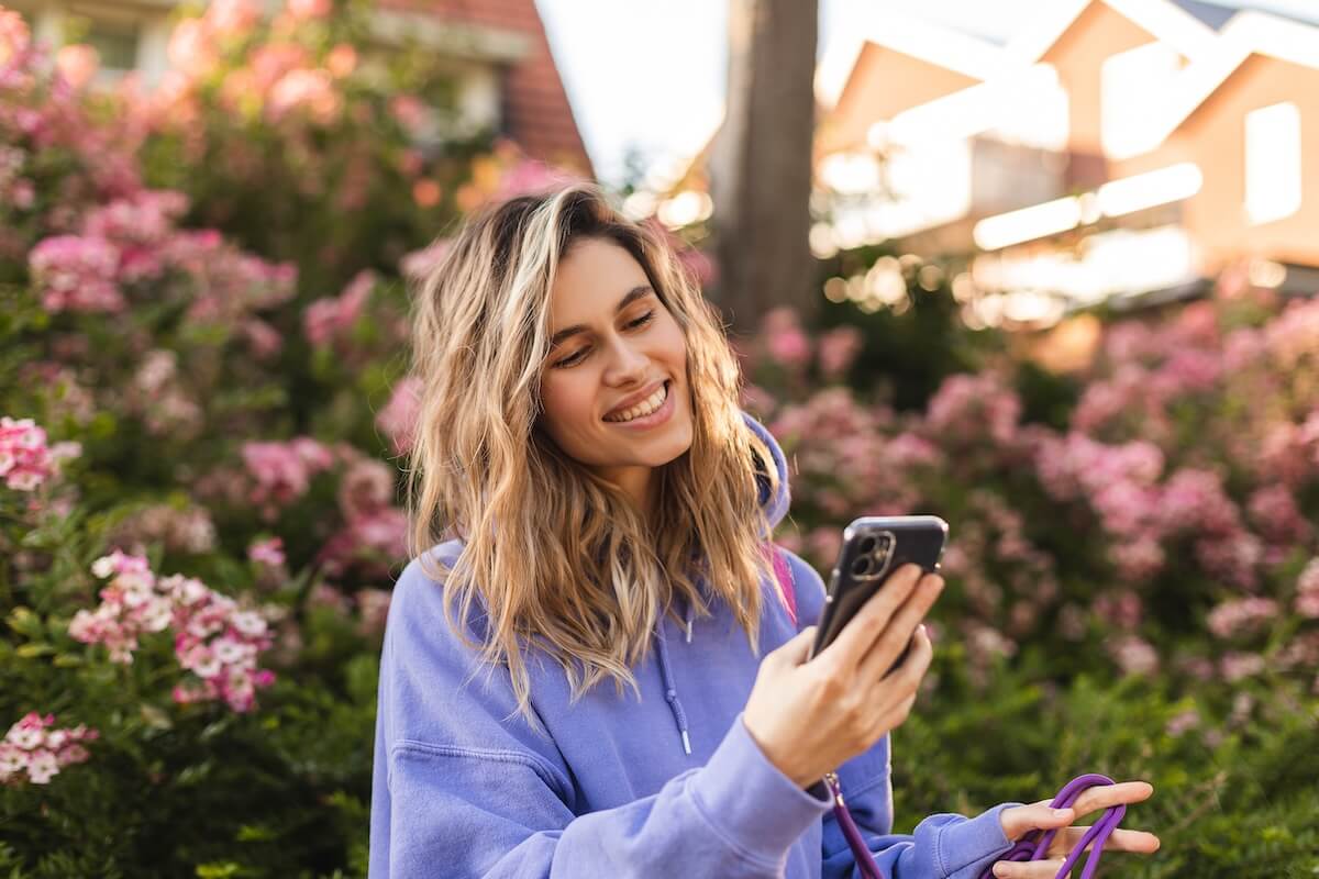 Shutterstock: Attractive blonde curly woman wear purple hoody hold mobile phone and chatting, messaging, look happy and smiling. Amazing enjoy woman walk near blossom flowers.