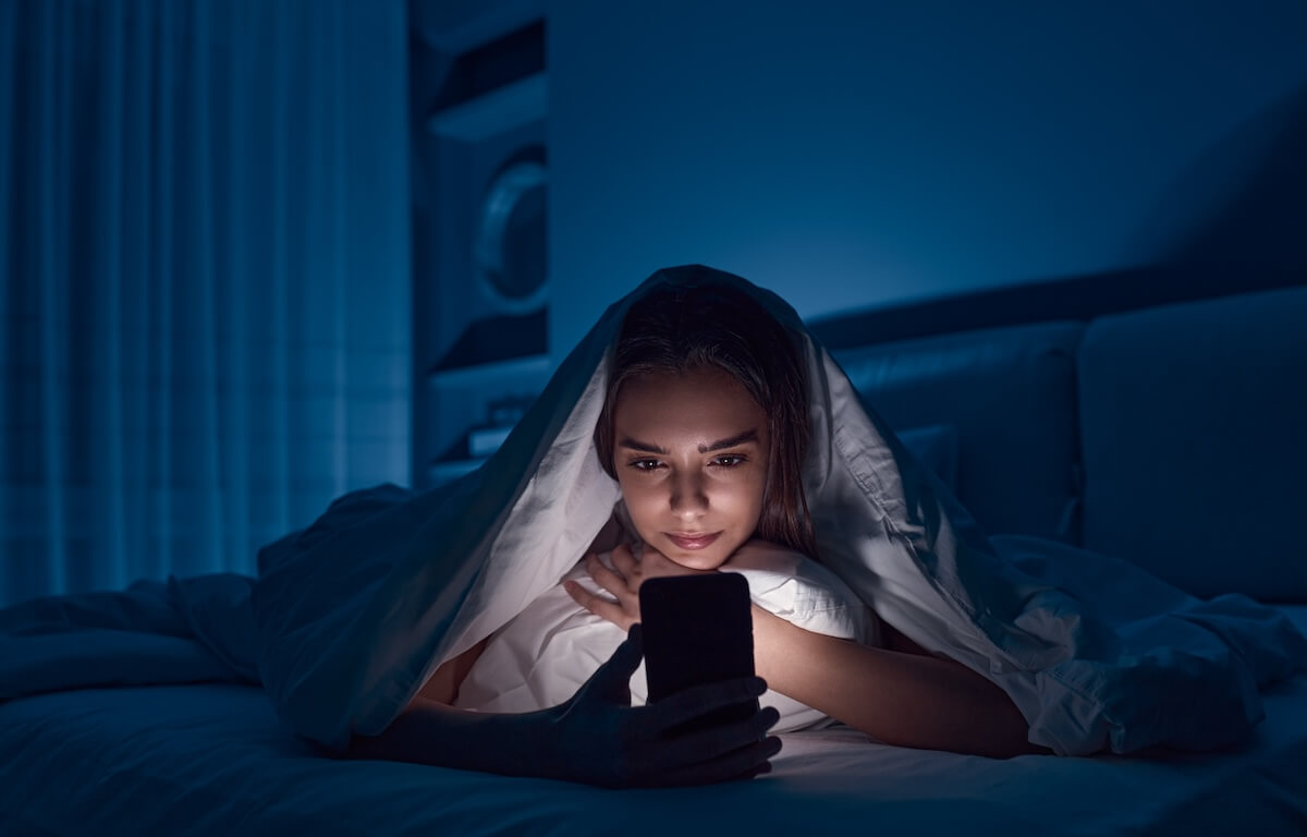 Shutterstock: Young female lying under blanket on bed and watching horror movie on mobile phone in dark bedroom in late evening