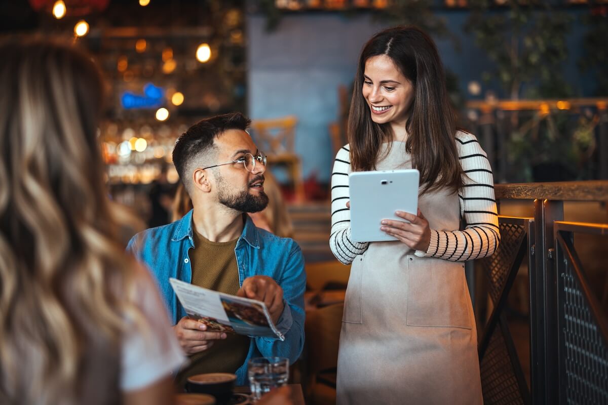 Shutterstock: A smiling waitress takes an order from guests at a cozy bar, with one man holding a menu and engaging in lively banter dressed in casual attire.