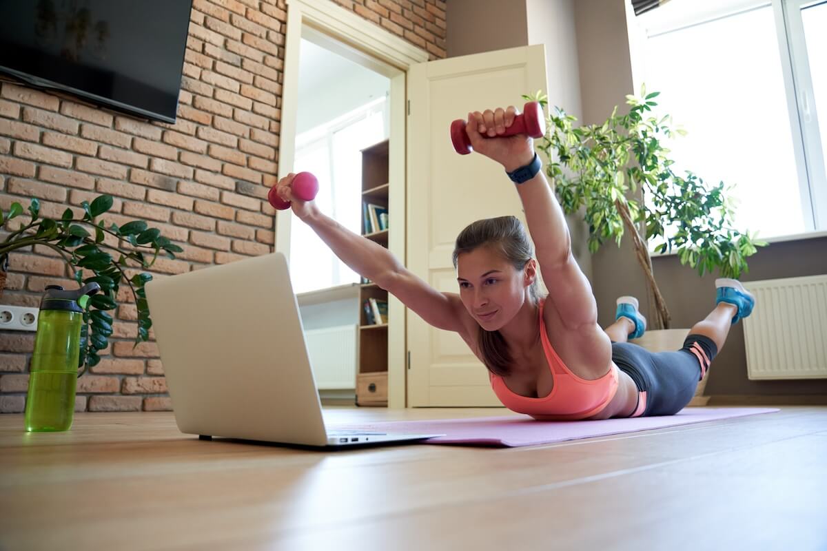 Shutterstock: Active fit sporty young woman holding dumbbells doing fitness exercise training abs and back at home on mat watching online video training tv tutorial sport live stream workout class on laptop.