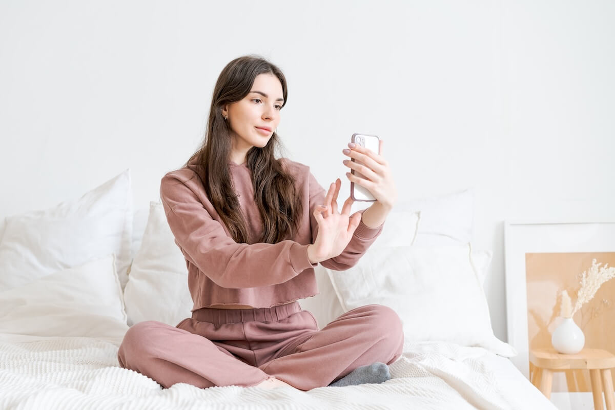 Shutterstock: Woman is sitting on a bed and taking a picture of herself with a cell phone. She is wearing a pink sweatshirt and pants
