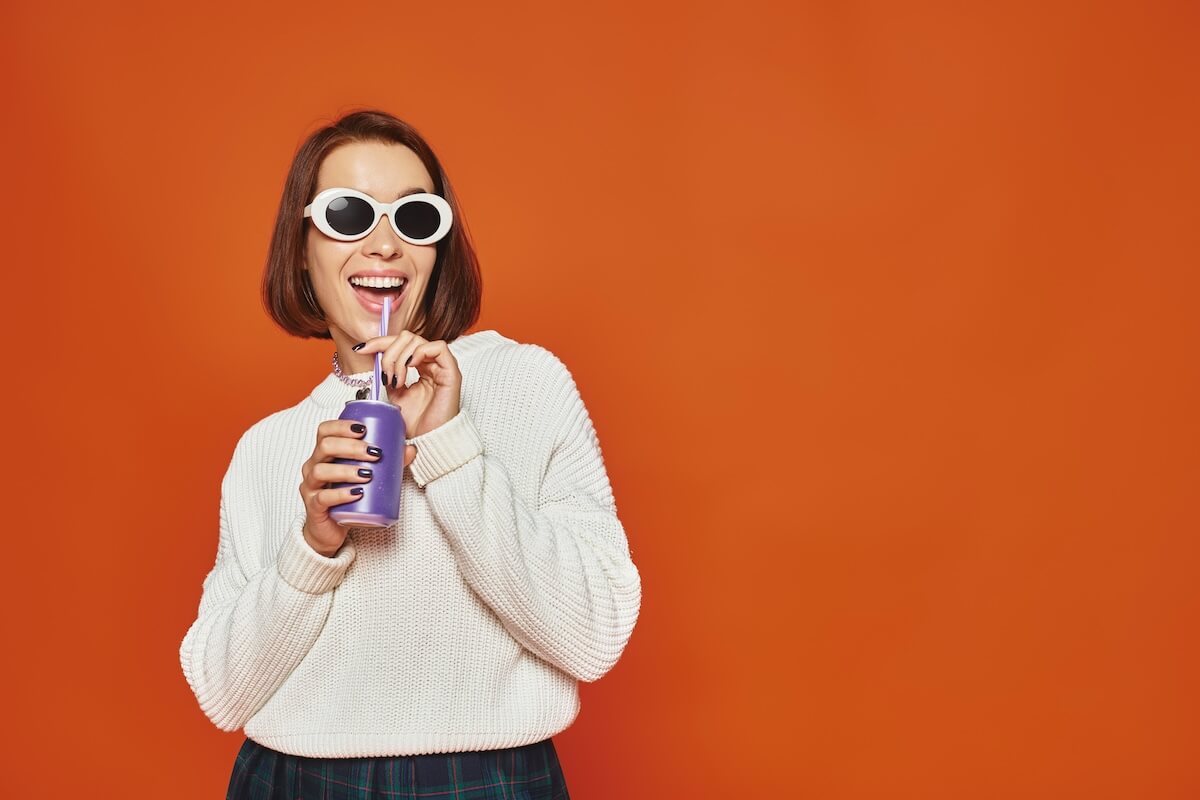 Shutterstock: Stylish young woman in white trendy sunglasses sipping soda drink from can on orange backdrop