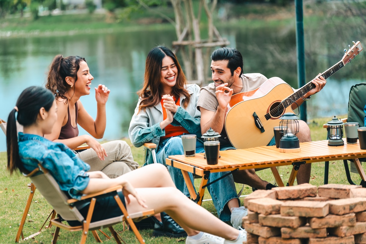 Shutterstock: A group of people are sitting around a table with a guitar and a fire pit. They are enjoying each other's company and having a good time