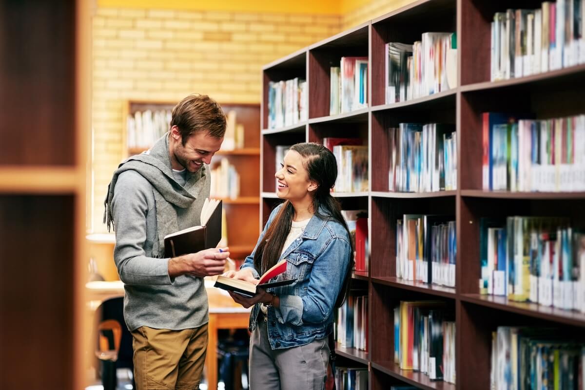 Shutterstock: People, students and library speaking with books, happy and campus for learning or university education. Conversation, academic scholarship and collaboration, exam preparation and college thesis
