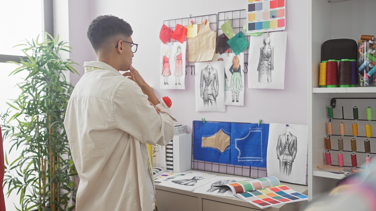 Shutterstock: A contemplative man in a tailor shop surrounded by fashion sketches and colorful thread spools.