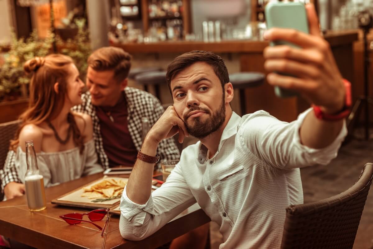 Shutterstock: Taking selfies. Upset bored sad pessimistic sorrowful blue dark-haired nice-looking bearded man in his 25s taking selfies while his friends hugging at the background