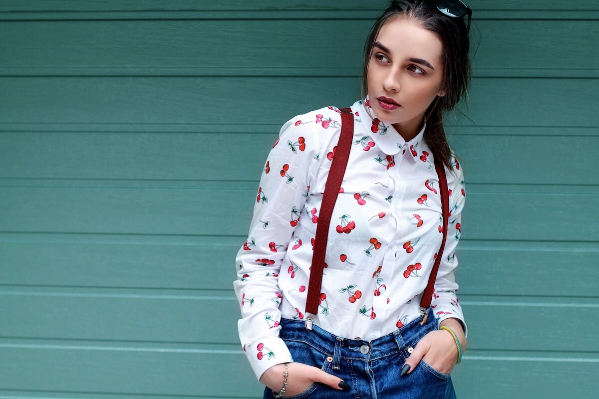 Shutterstock: Closeup fashion portrait of young pretty hipster girl with dark brown hairs wearing stylish ,denim braces for trousers glasses and shirt with funny print cherry .Mint bakground.Copy space