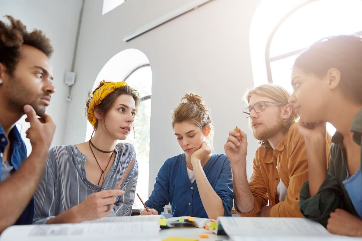 Shutterstock: Collaboration and brainstroming concept. Portrait of interracial friends gathering together sitting at table in classroom surrounded with books having debates expressing their opinions and views.