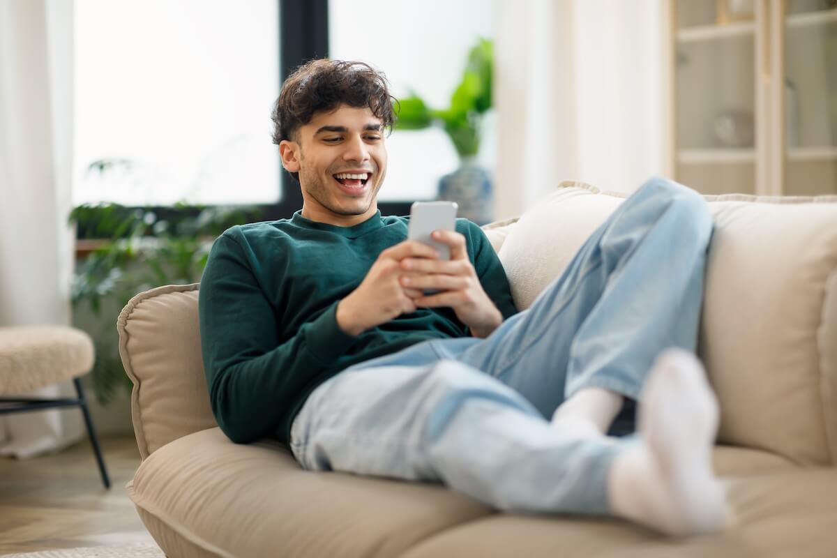 Shutterstock: Happy Middle Eastern Young Man Using Cellphone And Laughing Texting Lying On Sofa At Home. Cheerful Man Holding Smartphone Playing Game And Networking Online Having Fun On Weekend